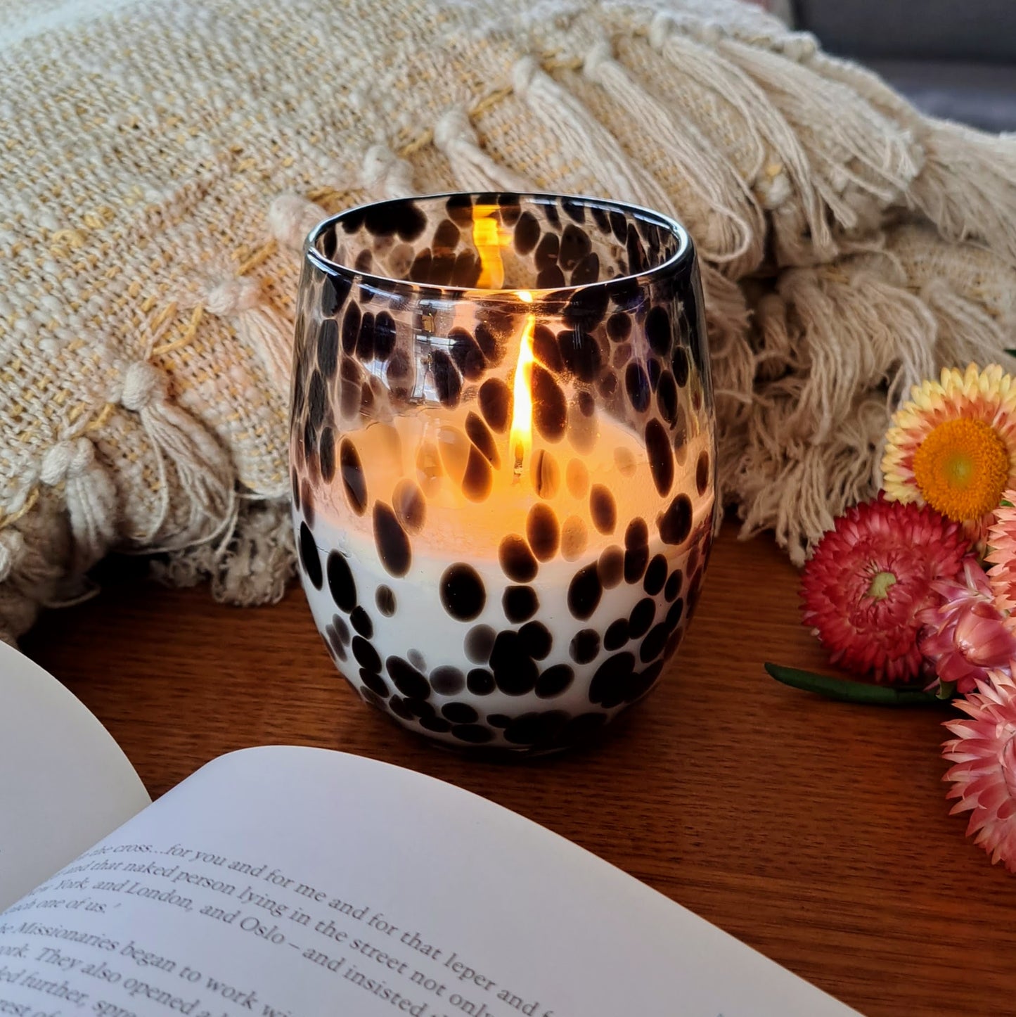 Lit black and white candle on a table next to flowers, a book and a rug